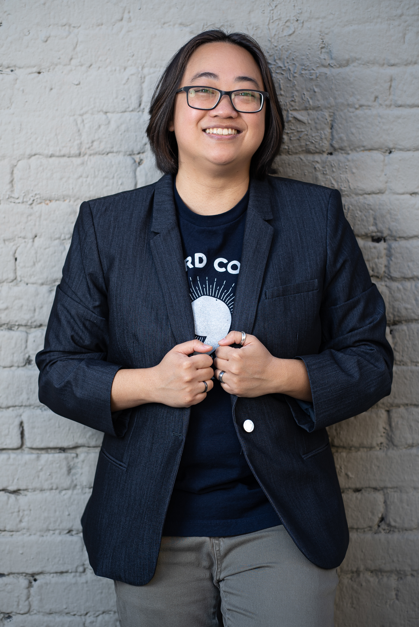 Photo of Stephanie Lim (Asian person with shoulder-length straight black hair), smiling and wearing black-rimmed glasses and a navy blue blazer with a dark blue "Oxford Comma" t-shirt underneath, standing outside in front of a grey brick wall.